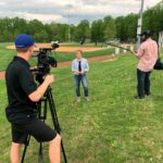 A cameraman is shown from behind filming a casually dressed woman near a baseball field as another film crew member holds a mic overhead.