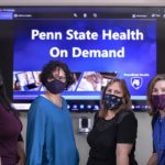 Four women wearing facemasks stand in front of a screen that reads Penn State Health OnDemand.