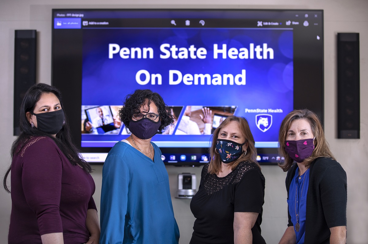 Four women wearing facemasks stand in front of a screen that reads Penn State Health OnDemand.