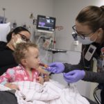 As young child sits on her mother’s lap in a hospital bed, a nurse administers a syringe of oral medicine to the pediatric patient.
