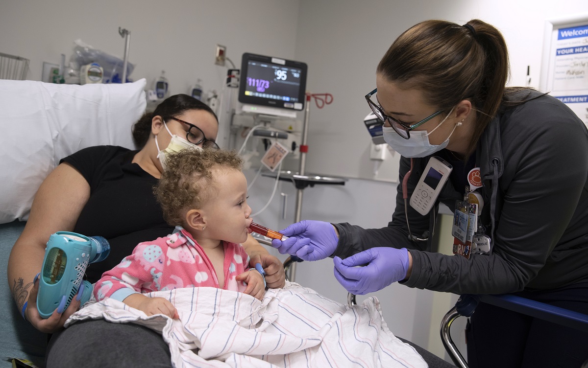 As young child sits on her mother’s lap in a hospital bed, a nurse administers a syringe of oral medicine to the pediatric patient.
