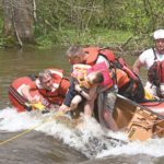 Rex Carmichael, wearing glasses and a life vest, saws tree roots to free a child in a creek as another paramedic holds onto the boy, who is slumped over his arm. Three other paramedics, wearing helmets and life jackets, sit in a river rescue boat.