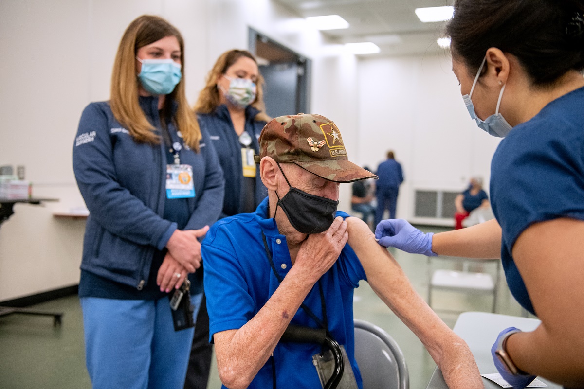 Robert Trate, wearing a mask and ball cap, rolls up his sleeve to receive a vaccination as registered nurse Judy Dee, wearing scrubs and a mask, leans toward him and cleans his bare upper arm. Physician Assistant Staci Gross Surgical Scheduler Jackelyn Ortega, wearing scrubs and masks, stand in the background.