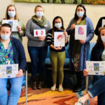 A group of eight people, six standing and two sitting, are pictured wearing face masks and holding signs.