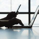 A man’s hand rests on a laptop computer. In his other hand he holds a cell phone.