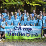 A group of teens and a few adults pose behind a large sign that reads “Camp Lionheart.” A building and some trees are in the background.