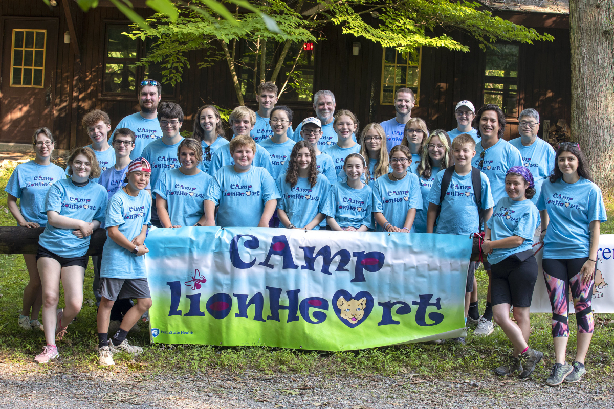 A group of teens and a few adults pose behind a large sign that reads “Camp Lionheart.” A building and some trees are in the background.
