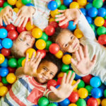 Three young children lie on their backs in a pit of colorful balls, their hands extended upward toward the camera.