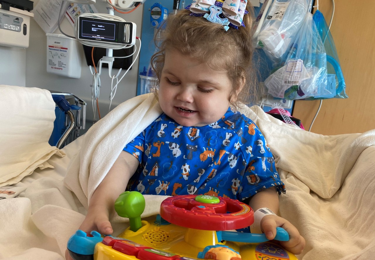 A young girl with a bow in her hair sits in a hospital bed with medical equipment behind her. She is playing with a plastic toy.