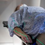 A person wearing a medical gown lays on a table. Lasers are visible on their body.