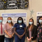 A man and four women stand in front of three research project posters hanging on the wall. Above the posters is a “Congrats to the Graduate” sign. All five people are wearing face masks.