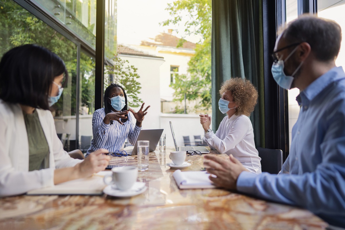 Diverse group of business people sitting together and having a meeting at the coffee shop. Brainstorming while wearing protective masks during a pandemic.