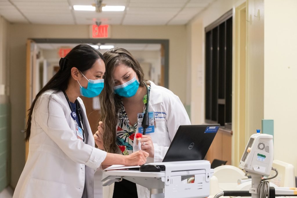 Two women in lab coats stand in a hospital hallway looking at a laptop together.