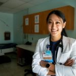 A smiling female doctor wearing a lab coat poses with her arms crossed in an exam room. She has long hair pulled back and is wearing a watch.