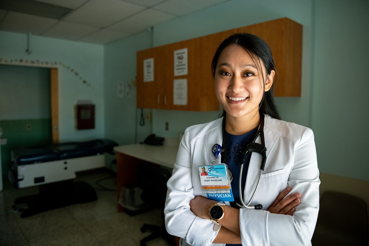 A smiling female doctor wearing a lab coat poses with her arms crossed in an exam room. She has long hair pulled back and is wearing a watch.