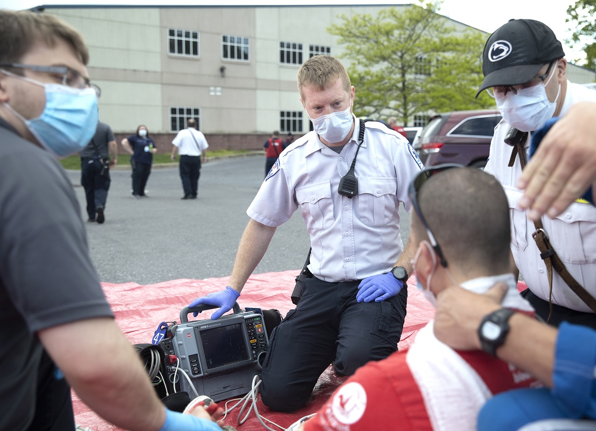 Four men are circled around a piece of equipment in a parking lot. They wear surgical masks. Two wear uniforms. Behind them other men and women in uniform stand in the parking lot.