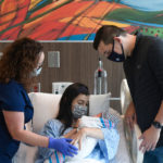 Two new parents smile at their newborn in a hospital patient room while a healthcare provider looks on.