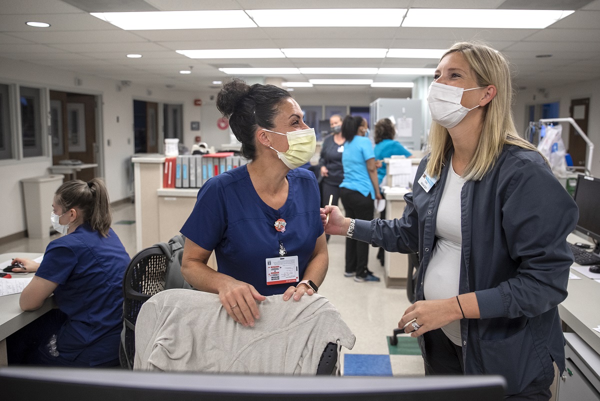 Two women in surgical masks and scrubs laugh at nurse’s station in a hospital hallway. Behind them, other health care workers cluster.