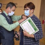 A man wearing mask, pants and vest, pins a small award pin to the jacket of woman wearing mask and business attire.