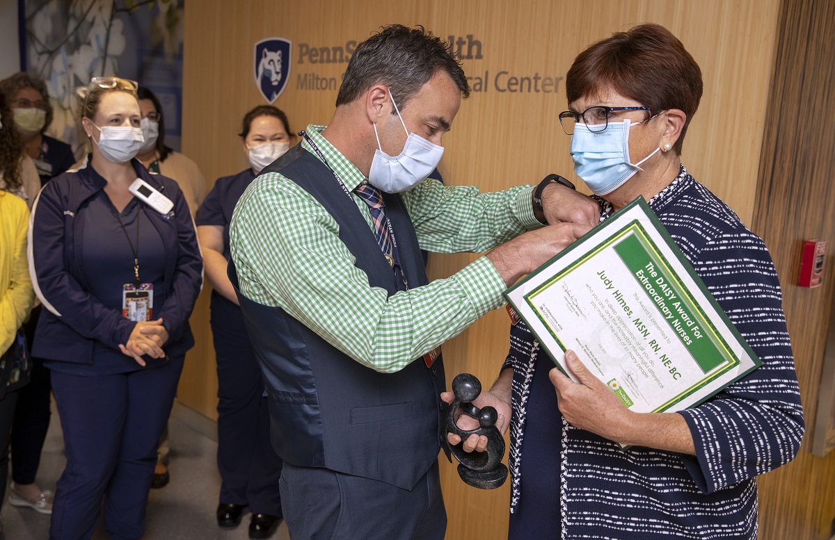 A man wearing mask, pants and vest, pins a small award pin to the jacket of woman wearing mask and business attire.