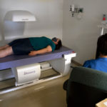 A woman participating in a clinical trial lays on a table in an exam room while a healthcare provider runs a test