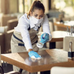 A worker in a cafeteria sprays and wipes down a small table. She is wearing a mask.