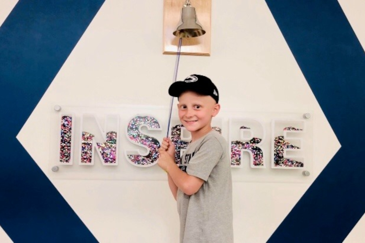 A boy wearing a baseball cap faces the camera and smiles, standing in front of a bell that hangs on a wall along with the word 