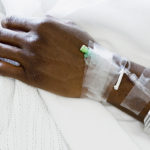 Close-up of a wrist and hand into which an IV line is inserted. Medical tape is on the arm, securing the line in place.