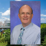 A head-and-shoulders professional photo of a man in a dress shirt and tie is superimposed on an aerial view of Penn State Health's campus in Hershey, Pennsylvania.