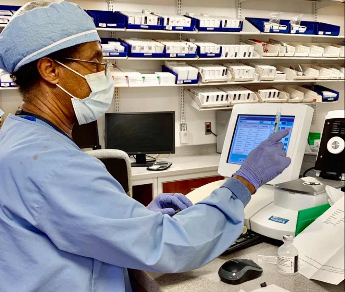 Tadesse Bogale touches a computer screen while holding a tube. He is wearing a face mask, scrubs, cap and surgical gloves. Behind him are shelves with medications.