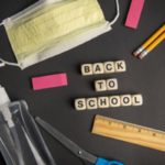 A variety of school supplies are spread out on a table. They include a pair of scissors, ruler, eraser, pencils, hand sanitizer, face mask, paper clips and colored pencils. Small wooden tiles spell out: Back to school.