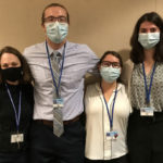 1 man and 3 women are standing in a line with their arms around each other's shoulders. They are all wearing face masks.