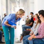 A woman in scrubs and a surgical mask stoops to speak with a woman and a child, both masked, in a hospital hallway.