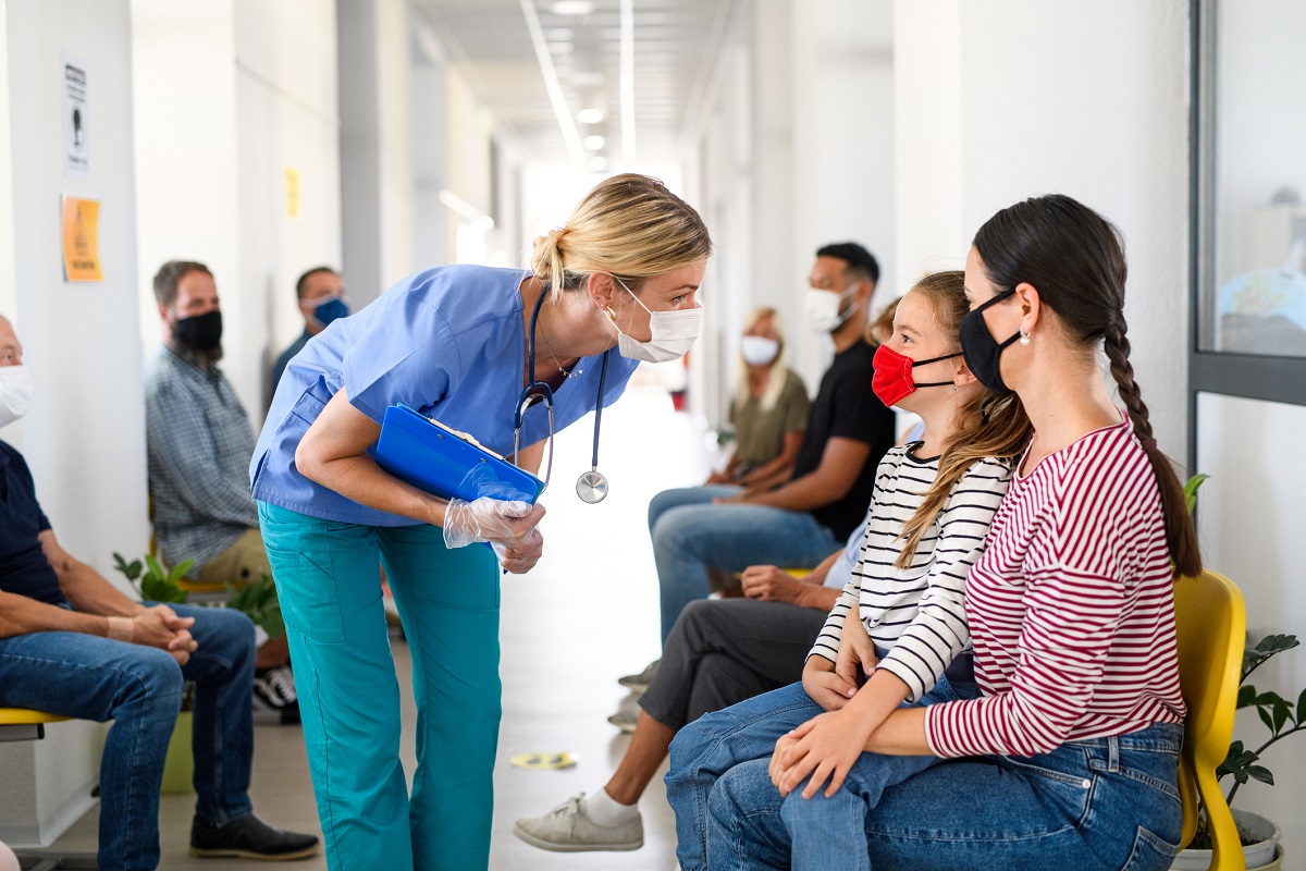 A woman in scrubs and a surgical mask stoops to speak with a woman and a child, both masked, in a hospital hallway.