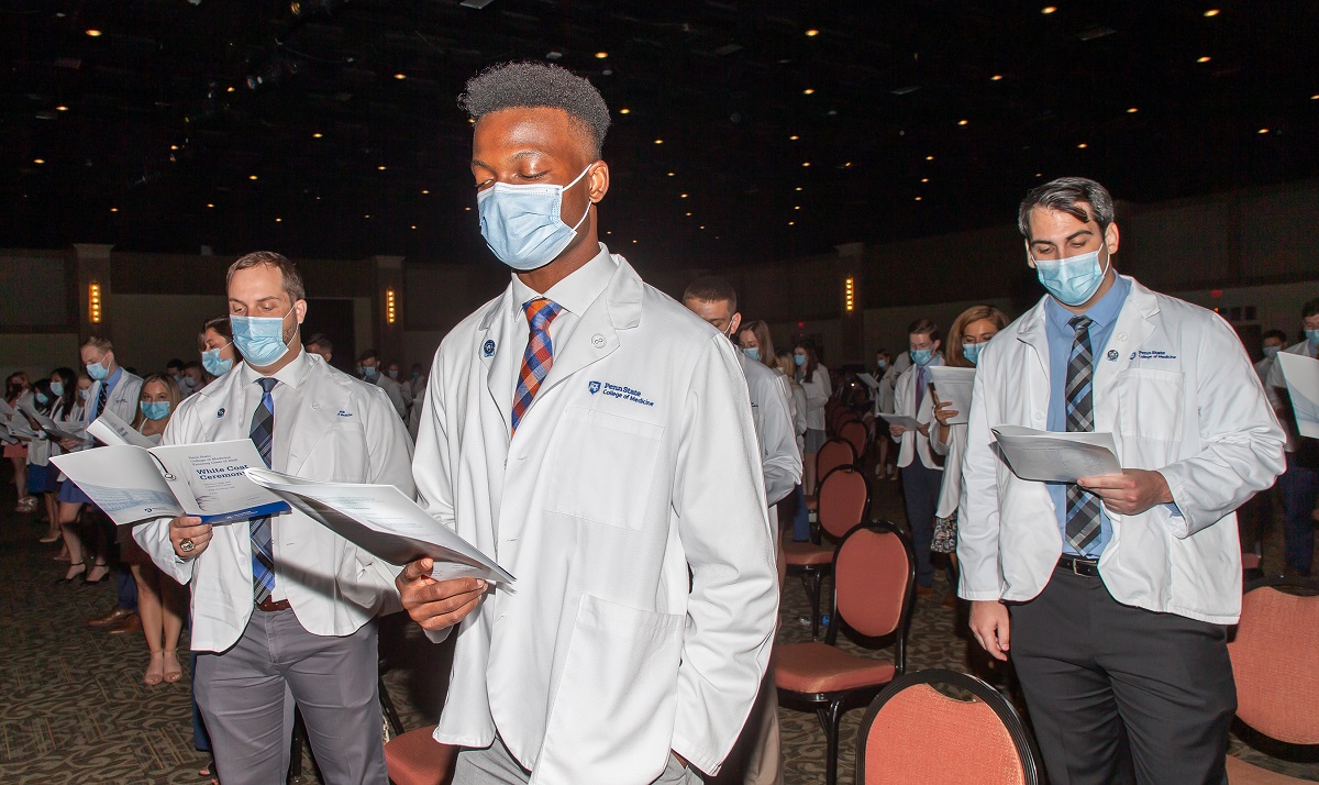 People in a crowded, but socially distanced, room, wear masks and white coats and look down at booklets.