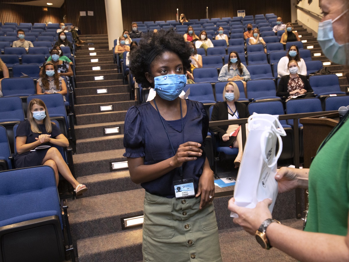 A woman wearing a surgical mask blouse and skirt raises a hand to take a bag from another woman at the front of an auditorium. In the seats behind them, others in masks look on.