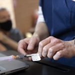 A close-up shows a woman's hand, opening a vaccination needle. In the background is a blurred, older man with a mask on, holding his sleeve up.