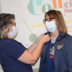 A woman in a mask pins something on the lapel of another woman in a mask.