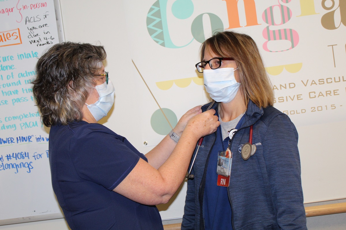 A woman in a mask pins something on the lapel of another woman in a mask.
