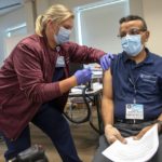 A woman wearing a face mask gives a COVID-19 vaccine to a man sitting in a chair and wearing eyeglasses and a face mask.