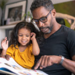 A man holds his young daughter on his lap as they both look down at a picture storybook. A bookshelf, artwork and plants are in the background.