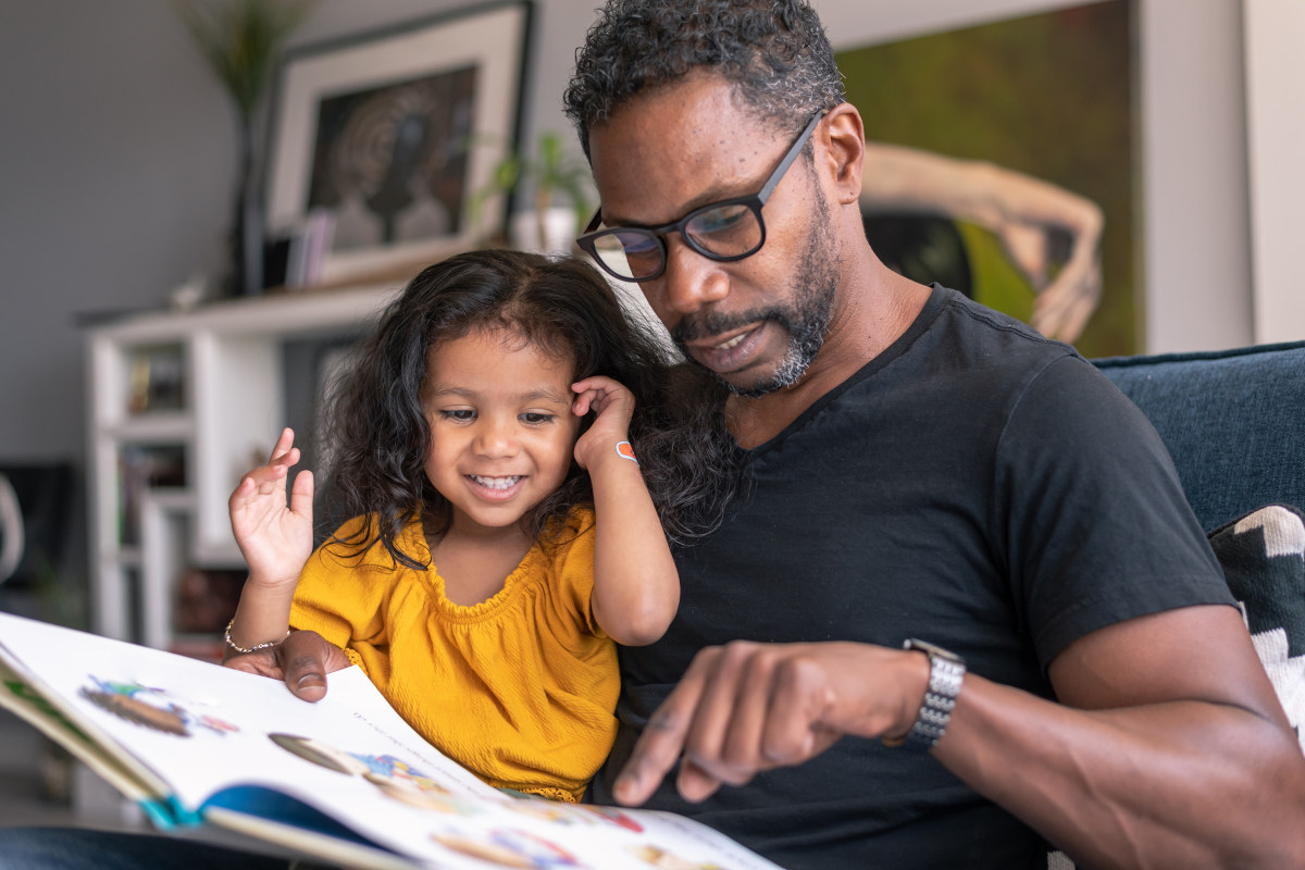 A man holds his young daughter on his lap as they both look down at a picture storybook. A bookshelf, artwork and plants are in the background.