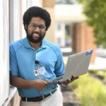 Penn State Health Information Services senior support specialist Sean Dolphin smiles as he leans against a wall outdoors. He is holding a laptop open. He has curly hair and a beard and is wearing glasses, a polo shirt and a nametag. Behind him are trees.