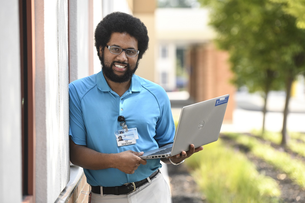 Penn State Health Information Services senior support specialist Sean Dolphin smiles as he leans against a wall outdoors. He is holding a laptop open. He has curly hair and a beard and is wearing glasses, a polo shirt and a nametag. Behind him are trees.
