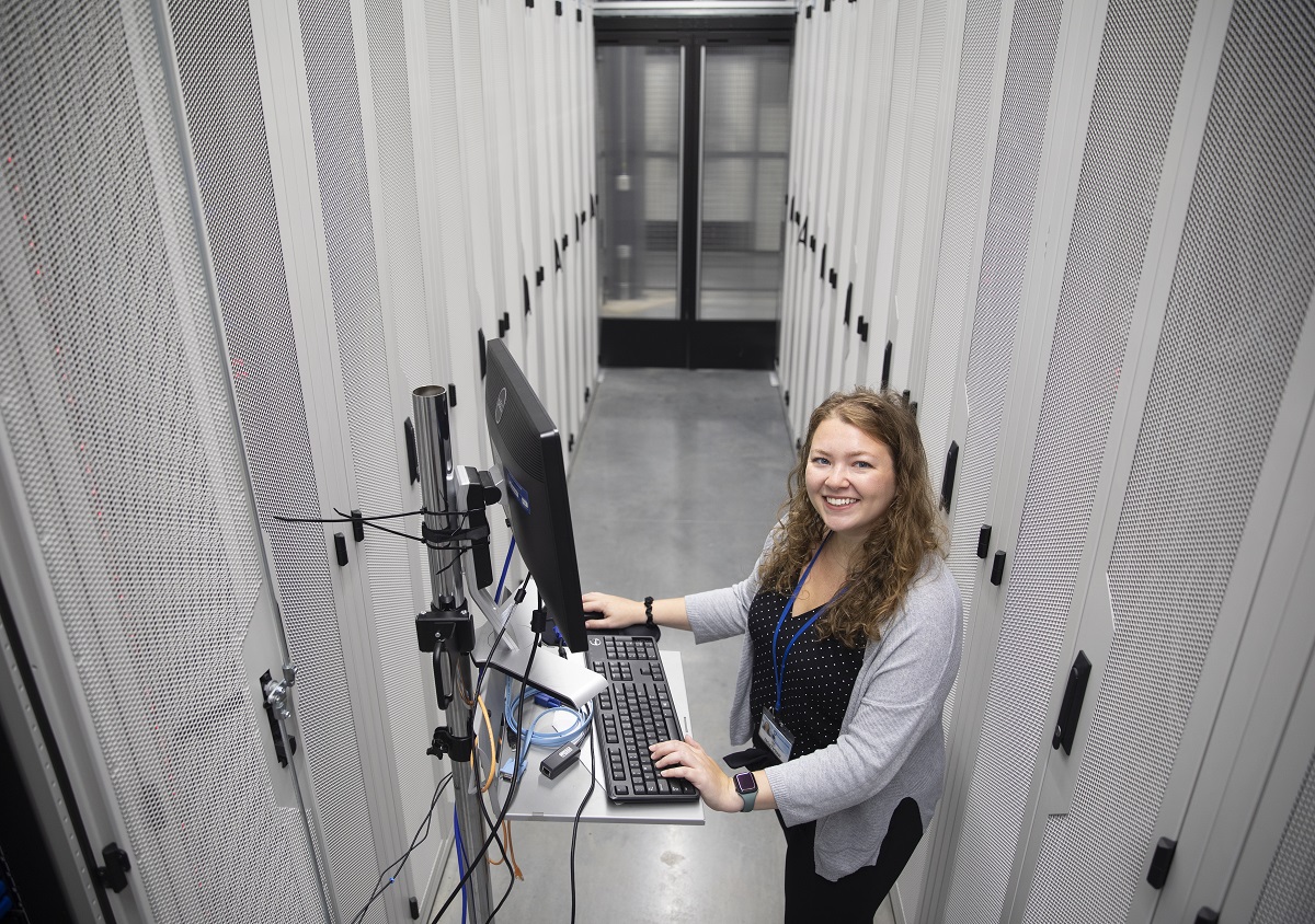 Lea Eller, a cyber defense infrastructure engineer at Penn State Health, stands in the middle of two rows of servers that are behind tall metal doors. She is standing at a laptop with her hands on the keyboard and looking up and smiling. She has long, curly hair and is wearing a top, long-sleeved sweater and nametag with a lanyard.