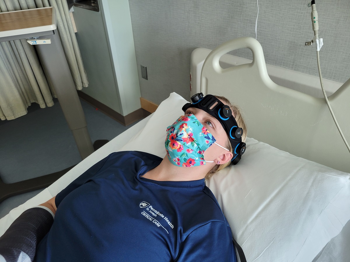 A woman wearing a face mask lays on an exam table to demonstrate the Ceribell Rapid Response Electroencephalogram system. She is wearing scrubs and a headband with integrated electrodes.