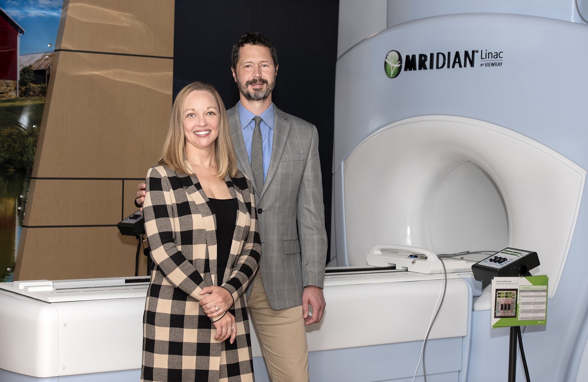 A man and woman stand in front of a Magnetic Resonance Imaging-guided linear accelerator.