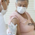 A woman wearing a mask receives an injection from a health care worker in gloves and wearing a mask.