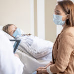 Woman wearing COVID mask in hospital bed with a woman who is also masked, seated beside her.