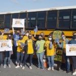 A group of 14 women stand in front of a school bus holding “Stuff the Bus” signs, pom-poms and foam fingers. Ten of them are wearing United Way T-shirts. Behind them is a Penn State Health pop-up tent.